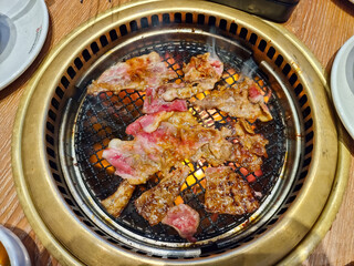 Top view of sizzling meat slices being grilled on a barbecue grill with glowing charcoal underneath, surrounded by plates on a wooden dining table, showcasing a delicious barbecue experience