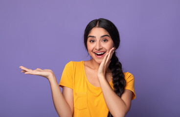 Wow, Look Here. Portarit of smiling young indian lady holding outstretched hand with open palm, advertising and showing free empty space and touching her cheek posing isolated on purple studio wall