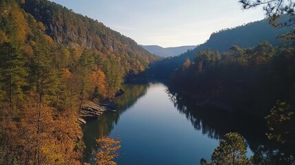 Fototapeta premium Serene autumn landscape calm river flowing through a valley surrounded by colorful trees and hills under a clear sky.