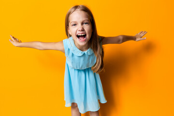 Joyful little girl in blue dress with arms outstretched against vibrant orange background,...