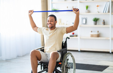 Happy black disabled man in wheelchair doing exercises with rubber band at home. African American...