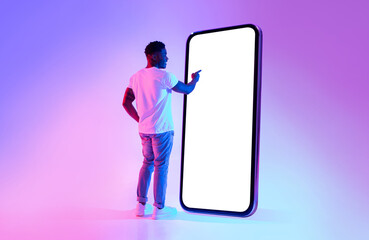 Full length of young black guy touching screen of big cellphone in neon light, mockup for mobile...