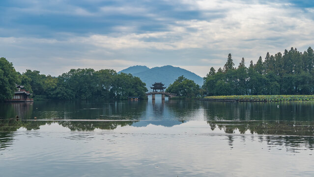 A beautiful calm lake. Green vegetation, lotus tickets near the shore. An arched bridge, a Chinese gazebo- pagoda in the distance. Reflection on the water. Mountains against the blue sky and clouds. 