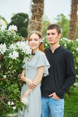 A young couple, a guy and a girl, are standing near a bush blooming with white flowers.