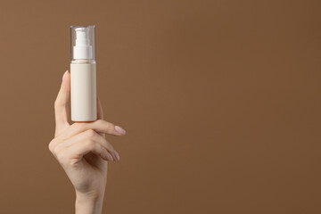 Woman's hand holds beige unmarked bottle for cosmetics. Mockup of container for serum, lotion, cream, retinol. Light brown background with copy space.