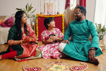 Family enjoying traditional Indian festival at home, smiling and sharing gifts while sitting on decorated floor