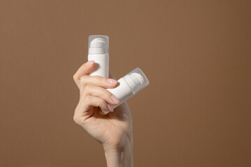 Woman's hand holds white unmarked bottles for cosmetics. Mock up of container for serum, lotion, cream, retinol. Light brown background with copy space