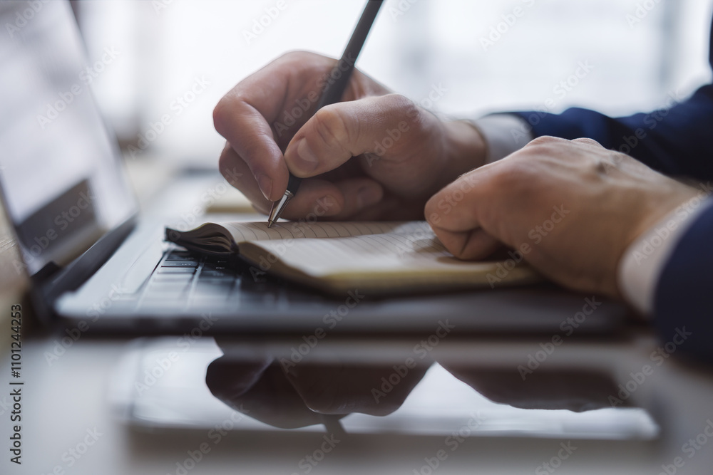 Aufkleber Close-up capture of a man's hand as he writes in a notepad on a sleek laptop, with an out-of-focus background
