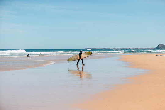A surfer walking on a sunny beach in portugal holding a surfboard