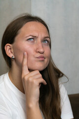 Young woman in a white t-shirt holds her chin with her index finger, squinting her eyes and...