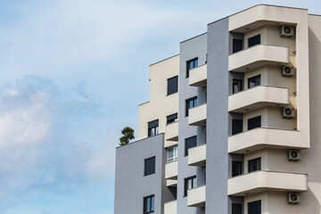 Modern apartment building with curved balconies and air conditioning units, featuring a minimalist design and a rooftop garden, under a blue sky