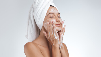 Beautiful woman with a towel wrapped around her head gently washing her face with foam. Ideal for...