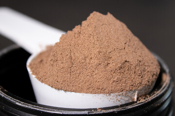 Close up of a scoop full with chocolate protein powder on a black can, grey table background
