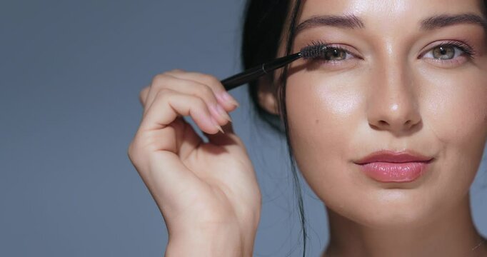 Beautiful woman with glowing skin and natural makeup grooming her eyelashes with a brush in a studio setting. Perfect for beauty tutorials, cosmetics promotions, and skincare campaigns. 