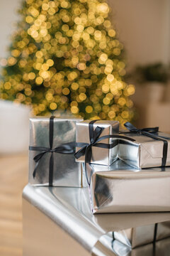 Close-up of a stack of Christmas gifts wrapped in silver paper in front of an illuminated Christmas tree