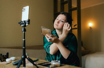 Plus size woman applying makeup on face with brush while recording vlog on smartphone for social media. Multiethnic fashion makeup artist vlogger live streaming makeup tutorial. Part of a series. UGC