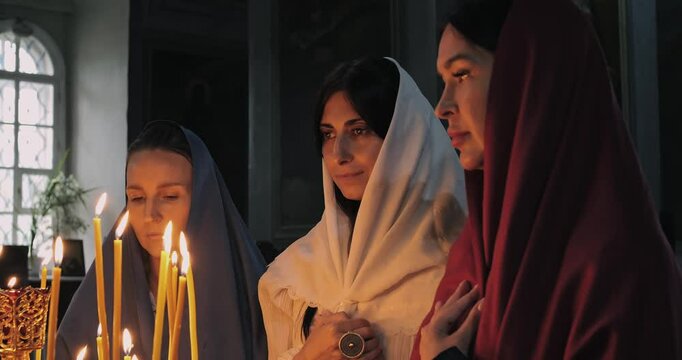 Three women pray in serene candlelit church, embodying devotion and spirituality. Holy atmosphere reflects Orthodox traditions and inner peace, illuminated by soft glow of candles and icons.