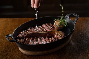A hand sprinkles salt over a sliced, medium-rare steak in a black cast iron pan with handles,...