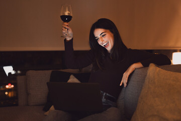 Happy millennial Caucasian woman sit on sofa celebrating with glass of wine in video call with friends having fun enjoying funny moment..