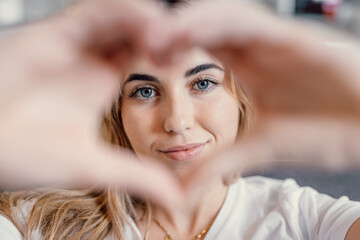 Fototapeta premium Beautiful girl showing heart sign with folded fingers smiling looking at camera, head shot portrait. Attractive optimistic young female demonstrate symbol of love feels happy posing alone at home.