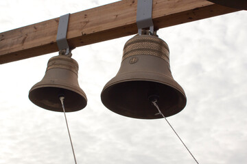 Two new bronze bells hang from a wooden beam against a cloudy sky. Bells in an Orthodox church.