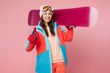 Snowboarder young woman wears warm blue padded jacket ski goggles mask spend extreme weekend winter season in mountains hold snowboard show thumb up isolated on plain pink background. Hobby concept.