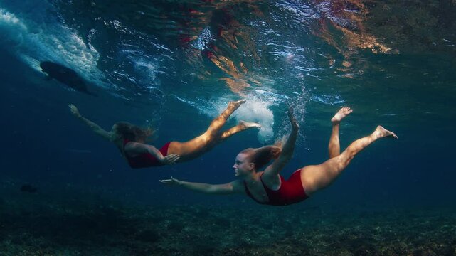 Two women in red suits dive under the breaking wave and swim confidently underwater