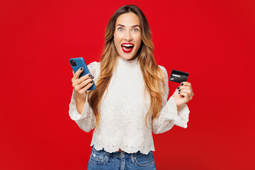 Young woman she wears white blouse casual clothes using mobile cell phone hold credit bank card doing online shopping order delivery booking tour isolated on plain red background. Lifestyle concept.