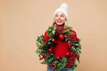 Merry young woman wear red warm cozy sweater hat posing hold in hand handmade wreath look aside...