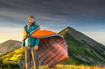 Stylish bearded man in a sleeping bag, fluttering in the wind like a cape, enjoying the sunrise in...
