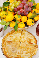 A plate with thin Russian pancakes and a bowl of fruit on the festive table.