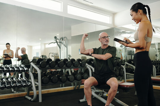 Athletic man lifting weights in modern gym while trainer with clipboard observes progress, reflecting dedication and guidance in fitness journey