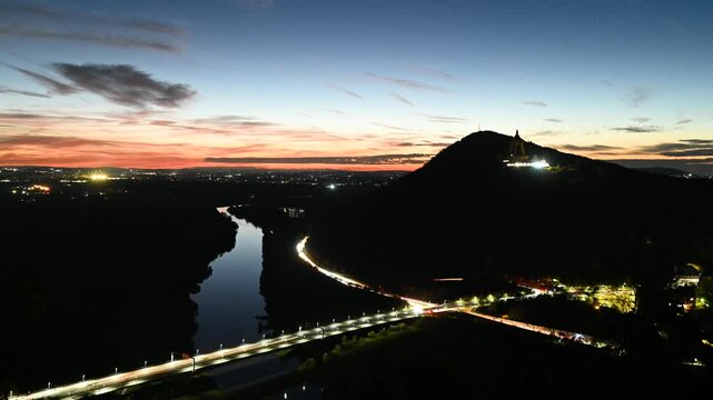 Time-lapse of Porta Westfalica during sunset with Kaiser Wilhelm Monument perched on a hill, vibrant city lights, and flowing traffic on a nearby bridge. 