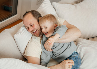 Dad hugs his son and they laugh merrily sitting on the beige couch in the living room