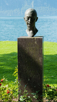A stone bust of Serge Rachmaninoff, situated by a lush green lakeside with distant mountains against a blue sky, immortalizes artistic legacy in natural harmony.