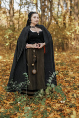 Young woman in medieval dress and cloak standing by tree in forest. Middle Ages