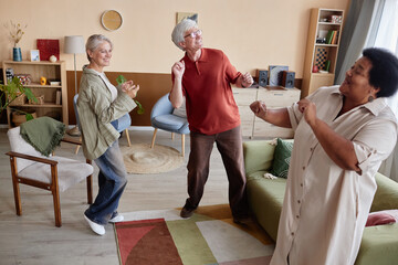 Full length view at group of joyful senior friends dancing together and listening to music enjoying entertainment in cozy living room