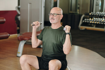 Senior man lifting dumbbells while sitting in gym setting. Focused expression showing commitment to health and fitness routine