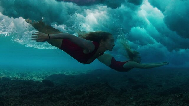 Two women in sensual red suits dive under the breaking wave and keep gliding underwater in the foam.