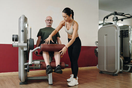 Female trainer instructing elderly man on leg exercise machine in fitness center. Senior receiving personalized guidance during strength training workout