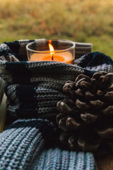 Orange scented wax candle in glass tumbler wrapped in a knitted scarf or sweater with a green natural background. Pine cone. Cozyness of slow living.
