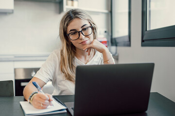 Millennial blonde girl sit at desk in living room study on laptop making notes, concentrated young woman work on computer write in notebook, take online course or training at home, education concept.