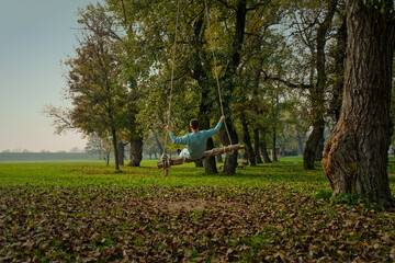 A men is sitting on a wooden swing suspended from a tree in a park. The scene is serene with lush...