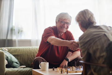 Portrait of grey haired senior man playing chess with wife or friend and smiling while shaking...