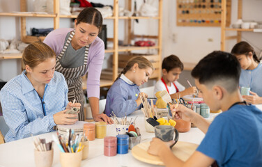 Children and teenagers sit at a table and paint ceramics at the workshop. Teacher shows how to...