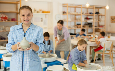 Teenage holds a clay product against the background of a teacher with children. Children working at the potter's wheel in a ceramics workshop