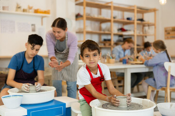 Boy in an apron is sculpting a mug on a potter's wheel. Child at a ceramic workshop with a teacher...