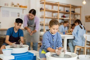 Positive children engaged in working at thrower's wheel in workshop together with teacher