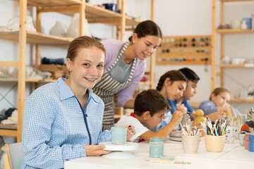 Positive teenage girl painting handmade ceramic cup with brush in pottery workshop