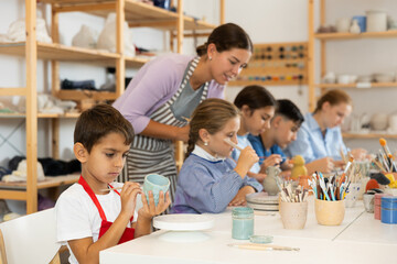 Attentive little boy painting handmade ceramic cup with brush in pottery workshop